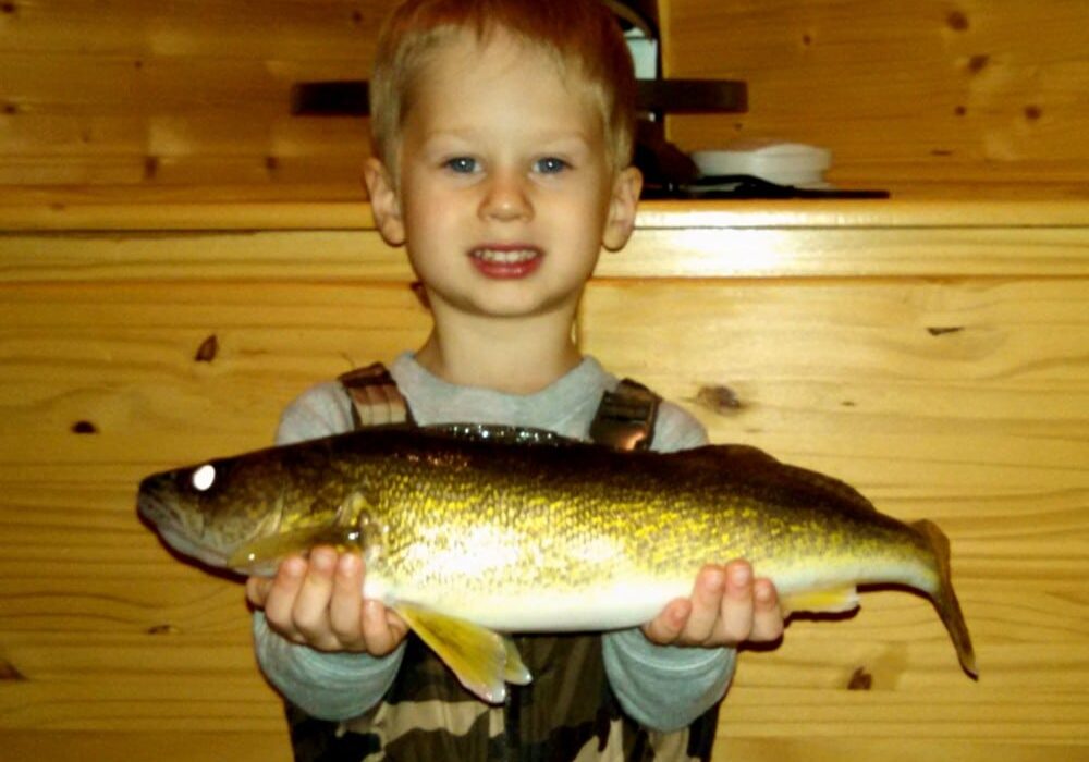 A young boy in camouflage overalls smiles while holding a large crappie with both hands in front of a wooden wall indoors.