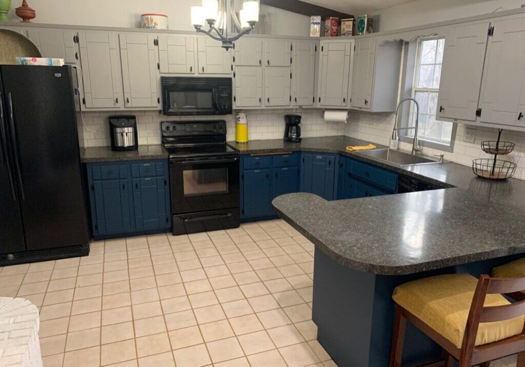 Modern kitchen with blue and gray cabinets, black appliances, a Crafters Edge black countertop, tiled floor, and a breakfast bar with two yellow-cushioned stools. A window above the sink lets in natural light.