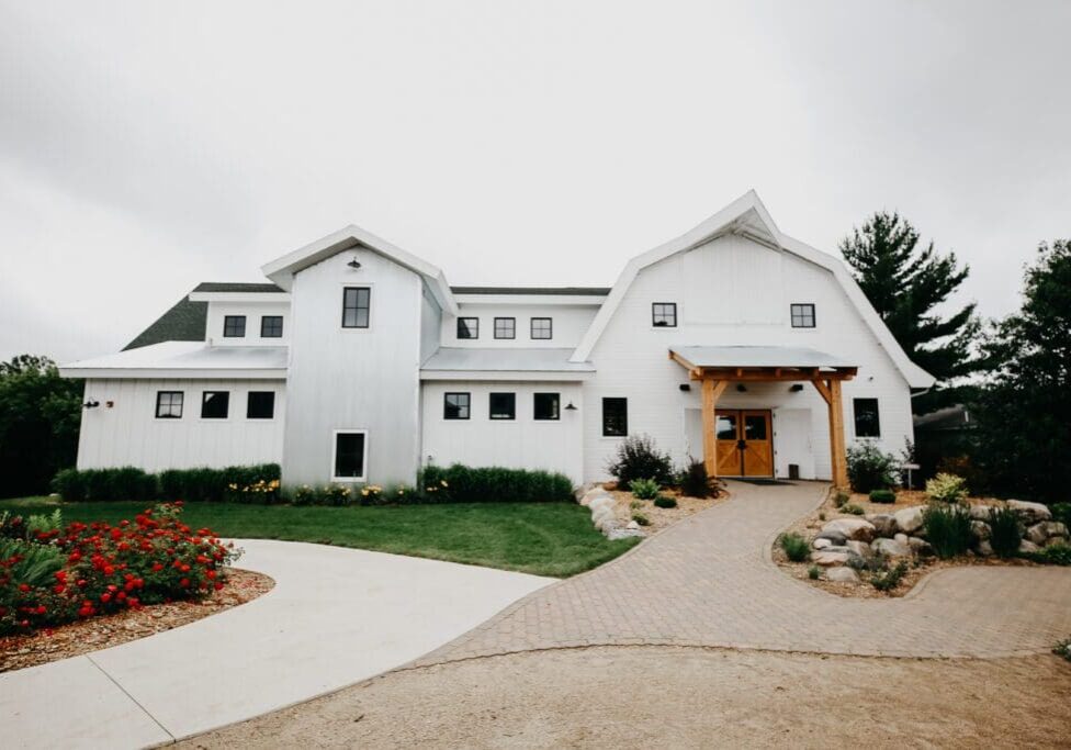 A large white modern barn-style building, Gathered Oaks features multiple gables and windows, surrounded by green grass, colorful flowerbeds, and a stone pathway leading to the entrance.