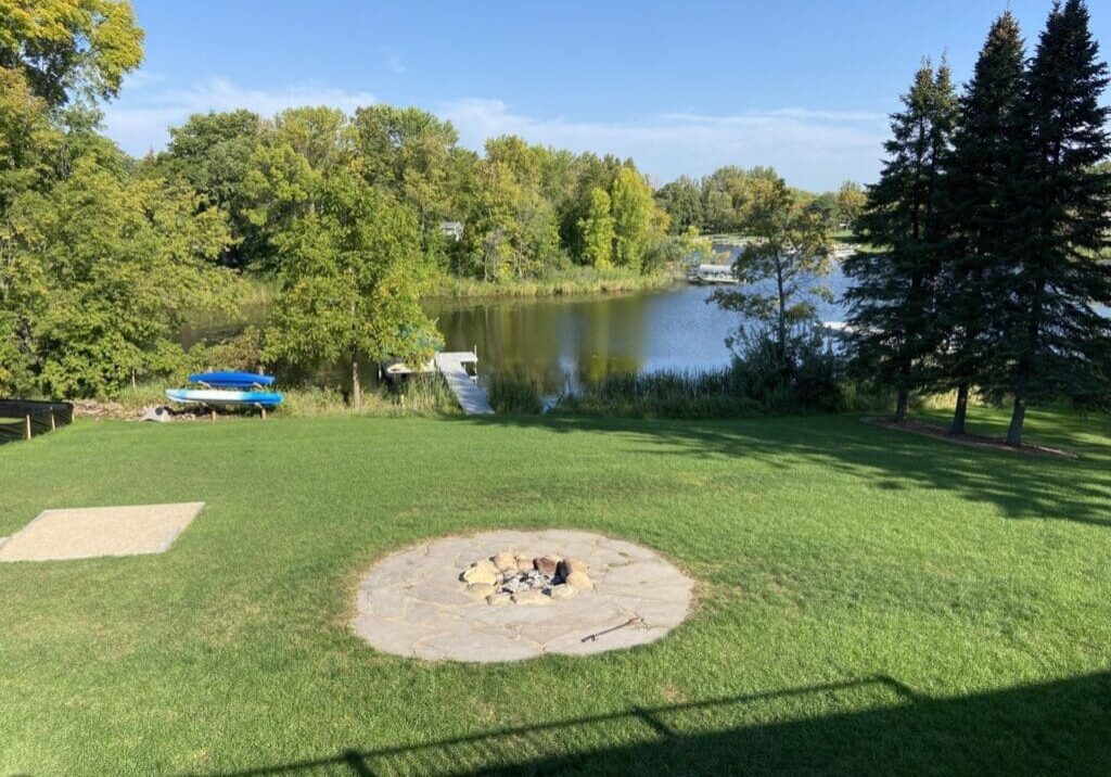 Lush green lawn with a stone fire pit in the foreground, a small lake and dock with boats in the middle ground, and trees surrounding Lake Le Homme Dieu under a clear blue sky near Crestwood Lodge.