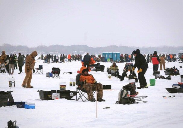 Hundreds of people bundled in winter clothing are taking on a fishing challenge, ice fishing on a frozen lake surrounded by gear, chairs, and portable shelters under a cloudy sky.