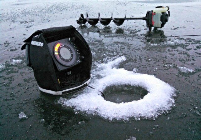 An ice fishing hole with a fish finder next to it sits on a frozen lake; an ice auger lies in the background on the icy surface, ready for action—perfect conditions described in the December 2014 ice fishing report.