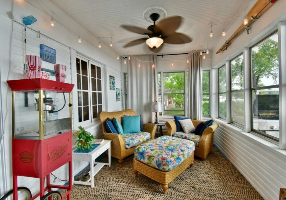 A cozy sunroom at Lake Le Homme Dieu Cottage- Mary's Place features wicker chairs and ottoman with blue cushions, a ceiling fan, string lights, popcorn machine, side table, and large windows with natural light and views—perfect for your Alexandria VRBO stay.