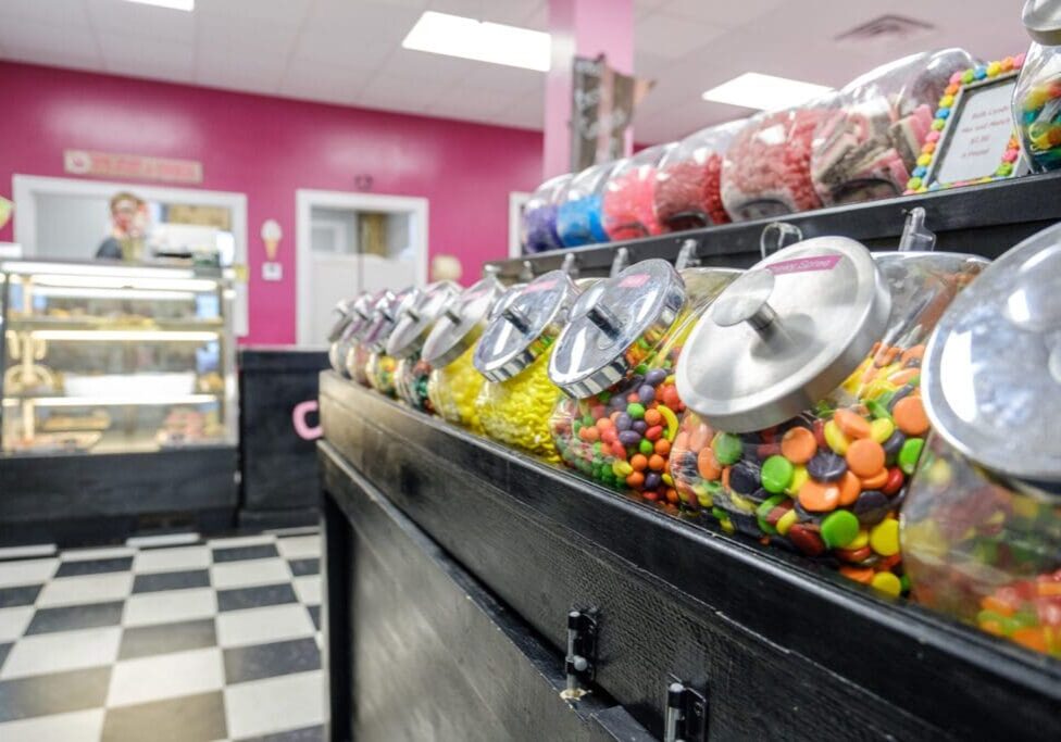 Clear jars filled with colorful candies line a black counter in The Sugar Shack, a bright candy shop with pink walls and checkered floors; a display case and person are visible in the background.