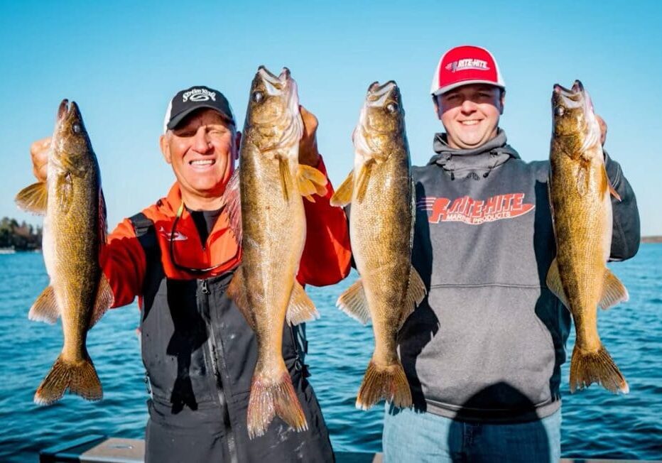 Two smiling men stand on a dock holding up four large walleye, each proudly displaying two fish. A blue lake and clear sky are in the background. The men wear casual outdoor clothing and caps, ready to share fishing tips from their successful day.