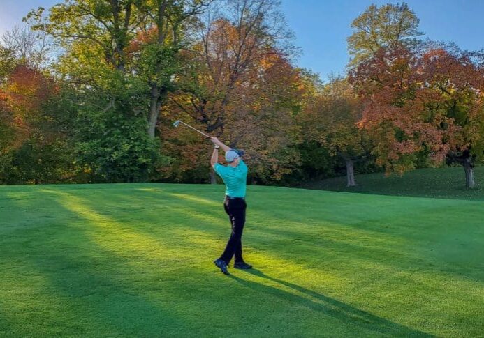 A golfer swings a club on a green course surrounded by colorful autumn trees in the late afternoon sunlight, savoring the peaceful scene as some golf courses are closing down.