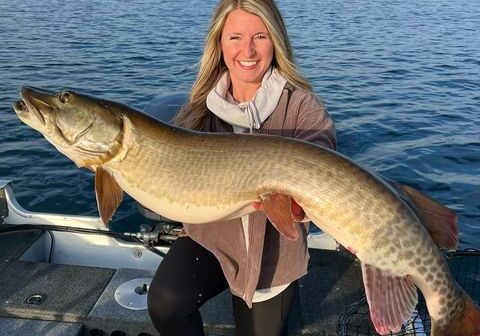 A smiling woman on a boat holds a large musky over her knees. The boat floats on a blue lake with a wooded shoreline in the background beneath a partly cloudy sky.