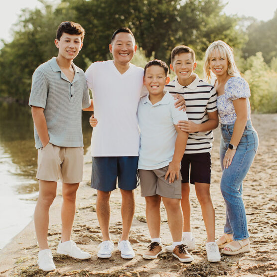 A family of five stands together on a sandy riverside, smiling at the camera. Trees and water are in the background, and everyone is dressed in casual summer clothes, ready for another day of adventure from the Summer Guide.