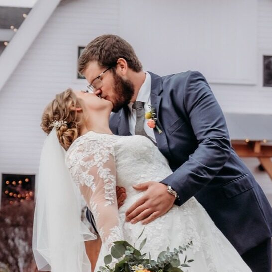 A bride and groom share a kiss outdoors. The bride wears a white lace gown and veil, holding a bouquet, while the groom is in a navy suit, gently dipping her. A white building is in the background.