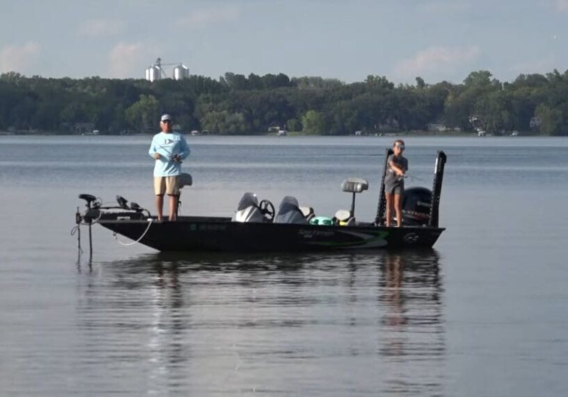 Two people are fishing on a black boat in a calm lake, surrounded by trees and distant buildings under a clear sky—an idyllic scene of fishing the Midwest.