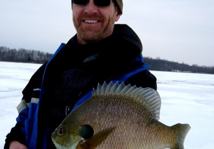 A man wearing sunglasses and a camouflage hat smiles while holding a trophy bluegill in front of him on a frozen, snowy lake. Trees are visible in the background under a cloudy sky.