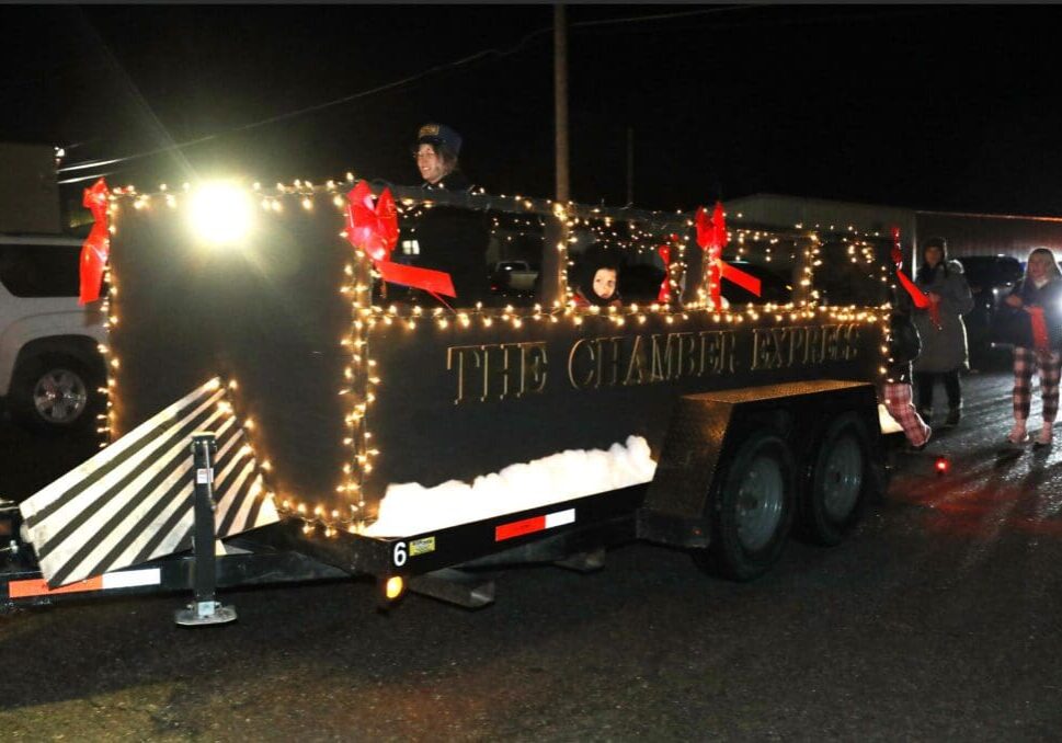 A parade float decorated as a black train car with "THE CHAMBER EXPRESS" in gold letters, festive lights, red bows, and people inside celebrates Christmas in the Fort as it moves down the street at night.