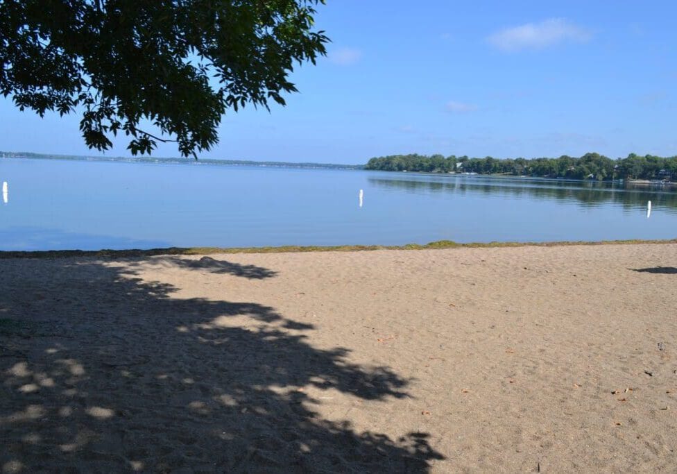 A sandy beach with some shadow from nearby trees overlooks the calm waters of Lake Le Homme Dieu Beach in Alexandria, MN. A clear blue sky stretches above, while white buoys float and a distant green treeline lines the far shore.
