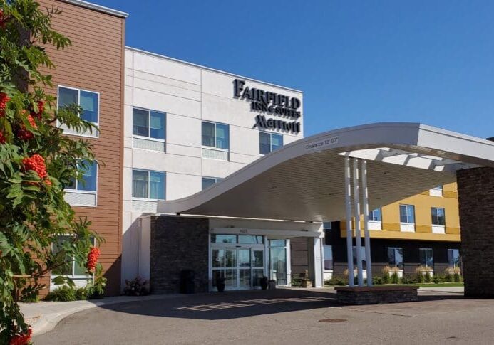 A modern Fairfield Inn & Suites Alexandria hotel entrance with a curved white canopy, stone pillars, large glass doors, and a tree with clusters of red berries in the foreground under a clear blue sky.