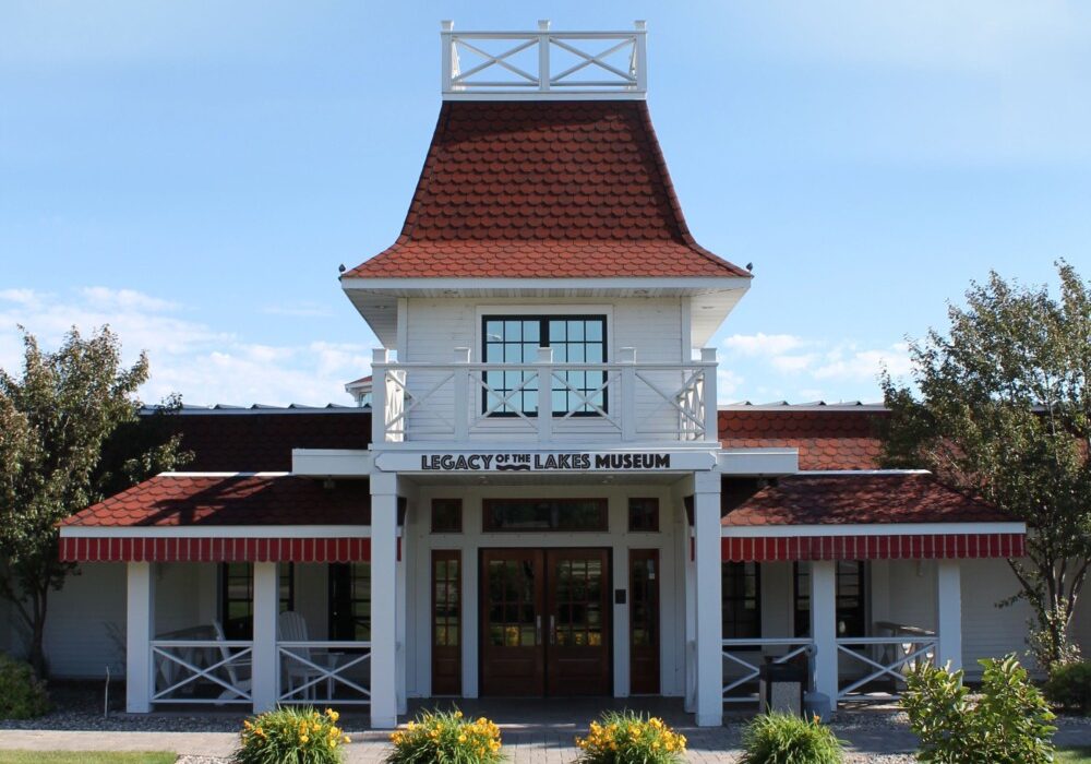 The Legacy of the Lakes Museum is housed in a white building with a red roof, featuring a central tower, front porch, and landscaping with yellow flowers and trees beneath a clear blue sky.
