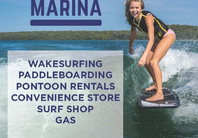 A young woman wakesurfs on a lake surrounded by trees near Alexandria, MN. Lake Carlos Marina offers lake country family fun with wakesurfing, paddleboarding, pontoon rentals, a store, surf shop, gas, and a phone number.