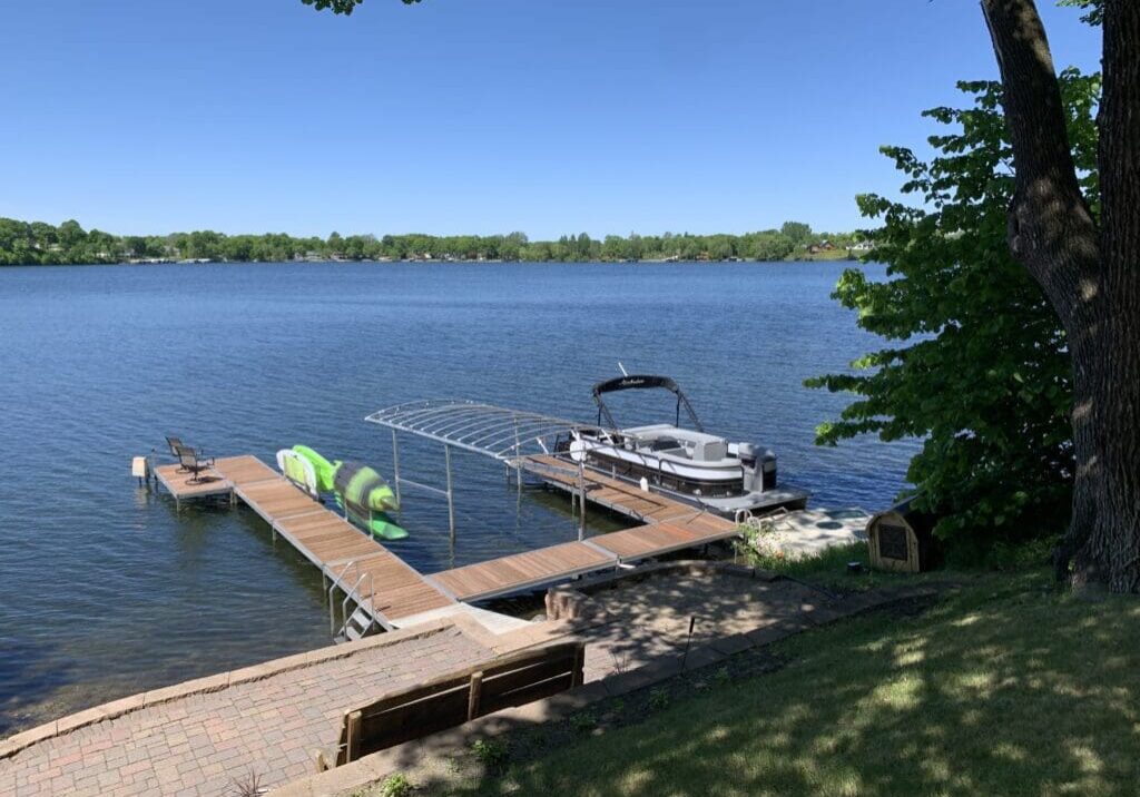 A peaceful lakeside scene at Lake Cowdry Retreat, with a wooden dock, pontoon boat, two green kayaks, and a bench on a grassy hill under a tree, all overlooking calm blue water and distant green trees beneath a clear sky.