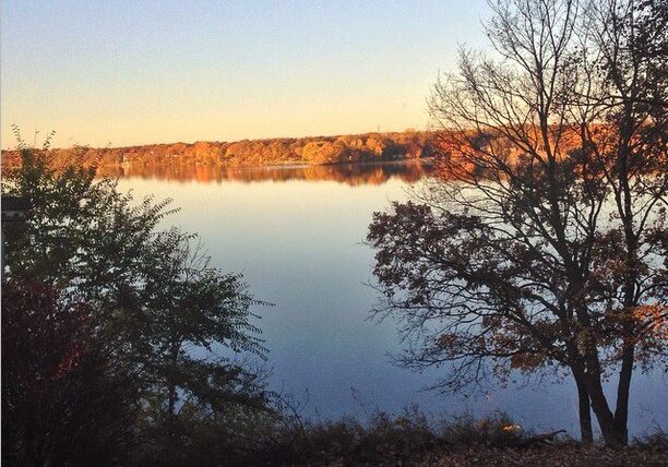 A calm Lake Geneva reflects trees with autumn foliage under a clear sky, with some bare and leafy trees in the foreground. Warm sunlight highlights golden and orange tones along the distant shore.