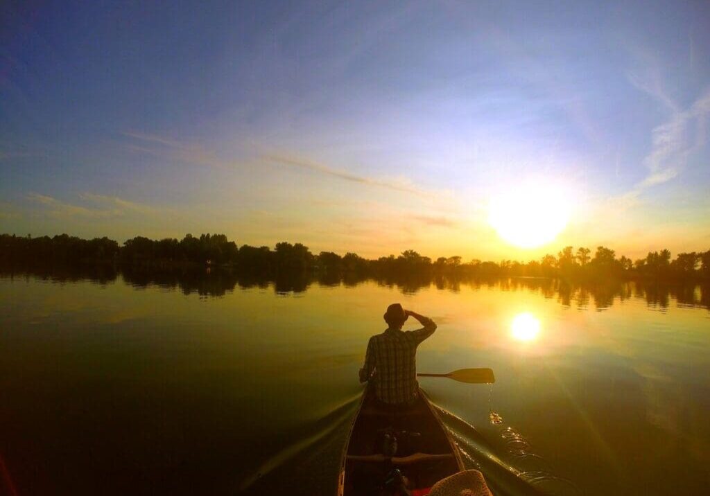 A person paddles a canoe on calm Lake Henry at sunset, with trees silhouetted along the shore and the sky glowing in shades of orange and blue.