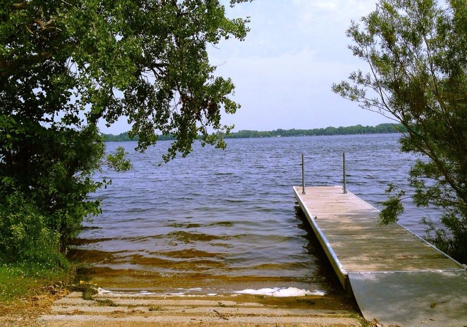 A small wooden dock extends into a calm lake reminiscent of Lake Louise, surrounded by trees and greenery under a partly cloudy sky. Steps lead down to the water’s edge, bordered by lush foliage on both sides.