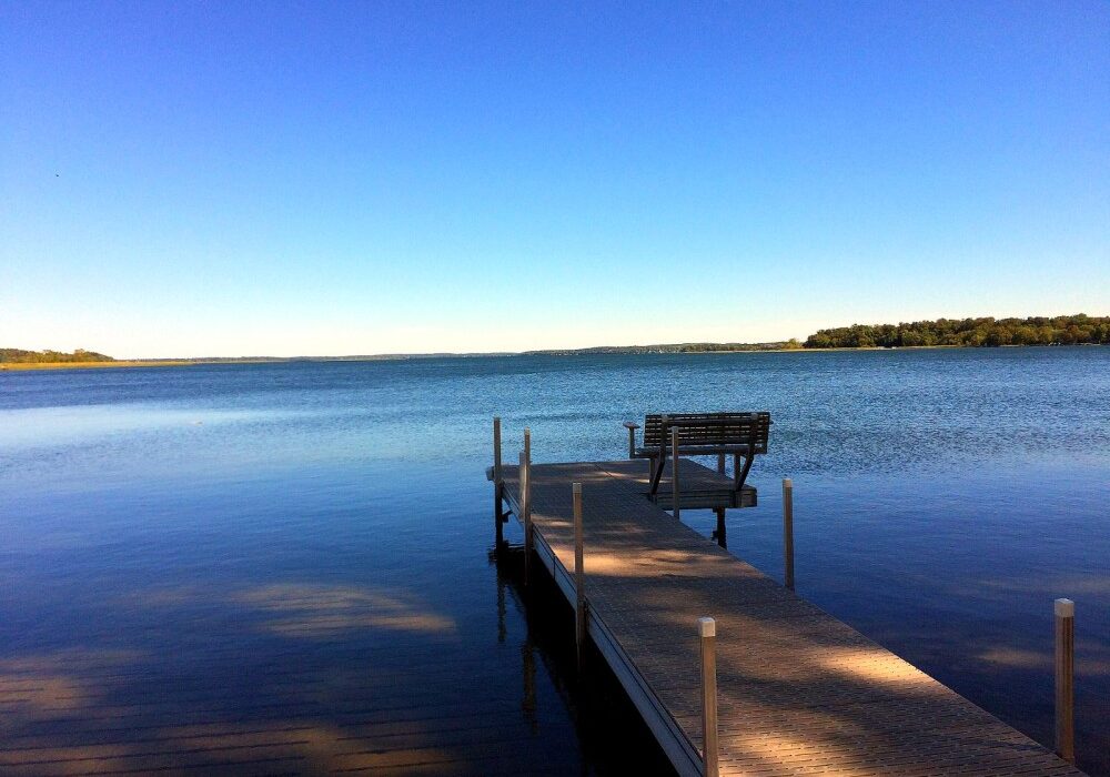A wooden dock with a bench extends over the calm blue waters of Lake Miltona, surrounded by distant tree-lined shores under a clear sky.
