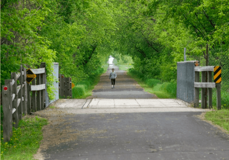 A person walks alone on the Lake Wobegon Trail, a paved path surrounded by lush green trees, with wooden and metal fences on either side and a bridge crossing under a canopy of foliage.