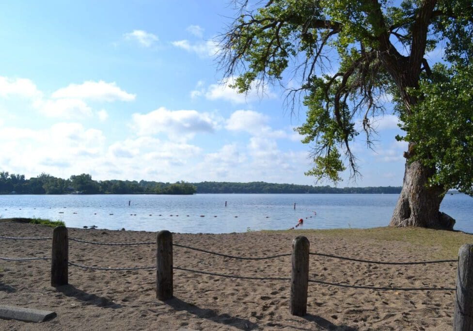 A sandy beach with wooden posts and rope fencing overlooks calm Lake Latoka in Alexandria, Minnesota. A large leafy tree stands on the right, and the sky is partly cloudy with sunlight shining through.