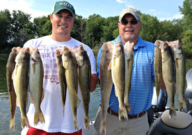 Two men standing outdoors on a boat in Alexandria, each holding up several large fish they caught, smiling at the camera. Trees and water are visible in the background, capturing a great moment from the August Fishing Report.