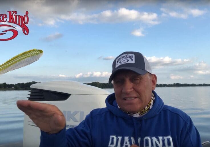 A man in a blue hoodie and Strike King cap, possibly Mike Frisch, gestures with his hand on a boat, with a Strike King logo and soft plastic fishing lure superimposed above his hand. A lake and cloudy sky serve as the backdrop.
