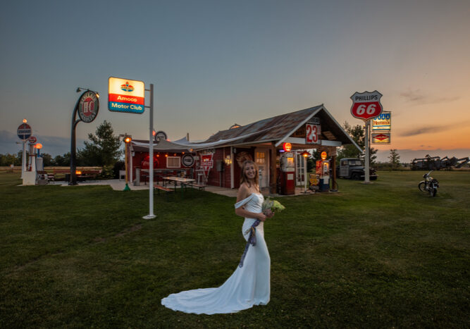 A bride in a white gown stands on grass at Adler Ranch in front of a vintage-style gas station at sunset. The red building, retro signs, and classic pumps set a nostalgic, Americana atmosphere.