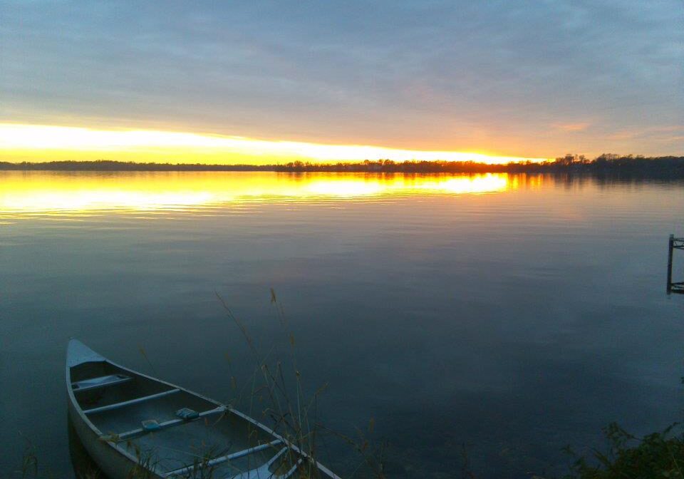 A canoe rests on the shore of Maple Lake at sunset, with golden sunlight reflecting off the calm water and a clouded sky overhead.