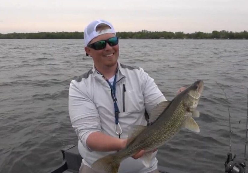 A smiling man wearing sunglasses and a white cap holds a large walleye on a boat in a lake, with shimmering water and distant trees in the background.
