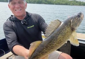 A smiling man sitting in a boat on a lake holds up a large walleye with both hands. Trees and water are visible in the background on a sunny day, capturing the perfect fishing moment in the Alexandria area.