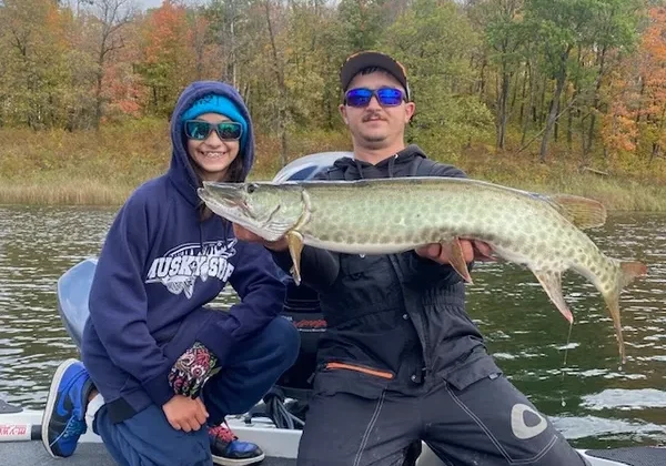Two people wearing sunglasses and hooded jackets on a boat, smiling and posing with a Mega Muskies catch. The background shows a lake and trees with vibrant autumn foliage.