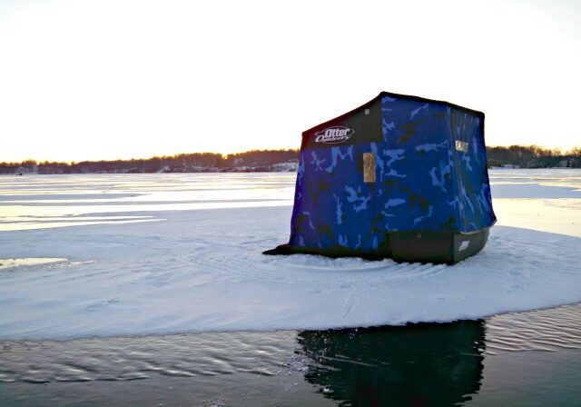 A blue ice fishing shelter sits on a snowy, frozen lake near an open area of water under a bright sky at sunset. Trees line the distant shore, creating a peaceful scene for fishing enthusiasts.