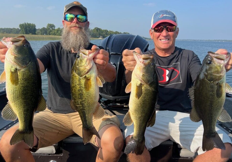 Two men sitting on a boat each hold up two large bass, smiling at the camera with a lake and trees in the background on a sunny summer day near Alexandria.