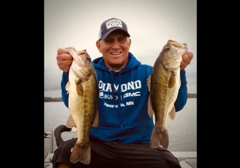 A smiling man in a blue hoodie and cap sits on a boat, holding up two large fish, one in each hand, ready to share his favorite fishing tips by the lake under a cloudy sky.