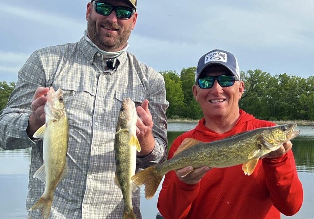 Two men in sunglasses and hats stand by a lake, smiling and holding up three fish they caught while fishing the Midwest. Trees and water are visible in the background under a cloudy sky.
