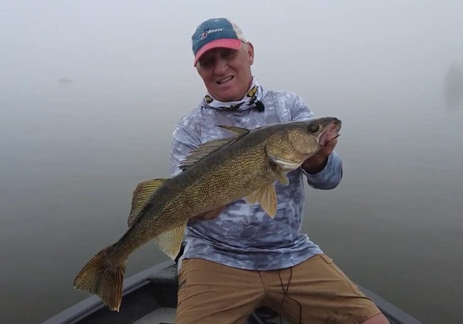 A smiling man in a cap and fishing attire sits in a boat on a foggy lake, proudly holding a large fish with both hands after catching walleyes as the weather cools down.