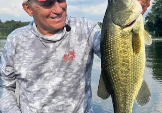 An older man in a cap and long-sleeve shirt smiles while holding a large fish beside fishing docks on a calm lake, with trees and a partly cloudy sky in the background.