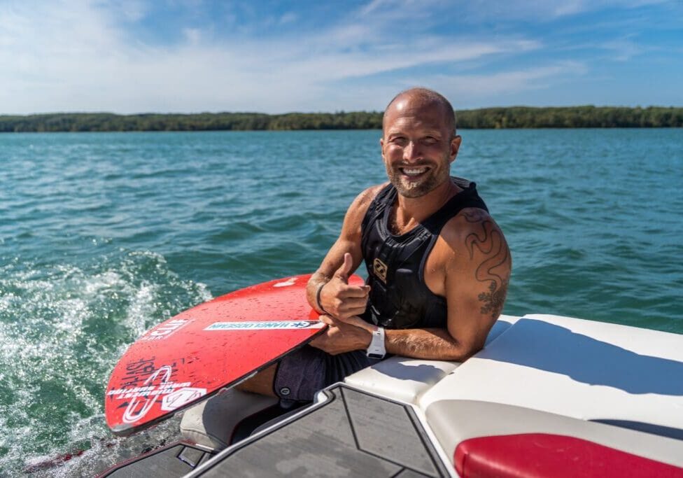 A smiling man in a black life vest sits on the edge of a boat holding a red wakeboard, ready for water sports, with blue water and a distant treeline in the background on a sunny day.