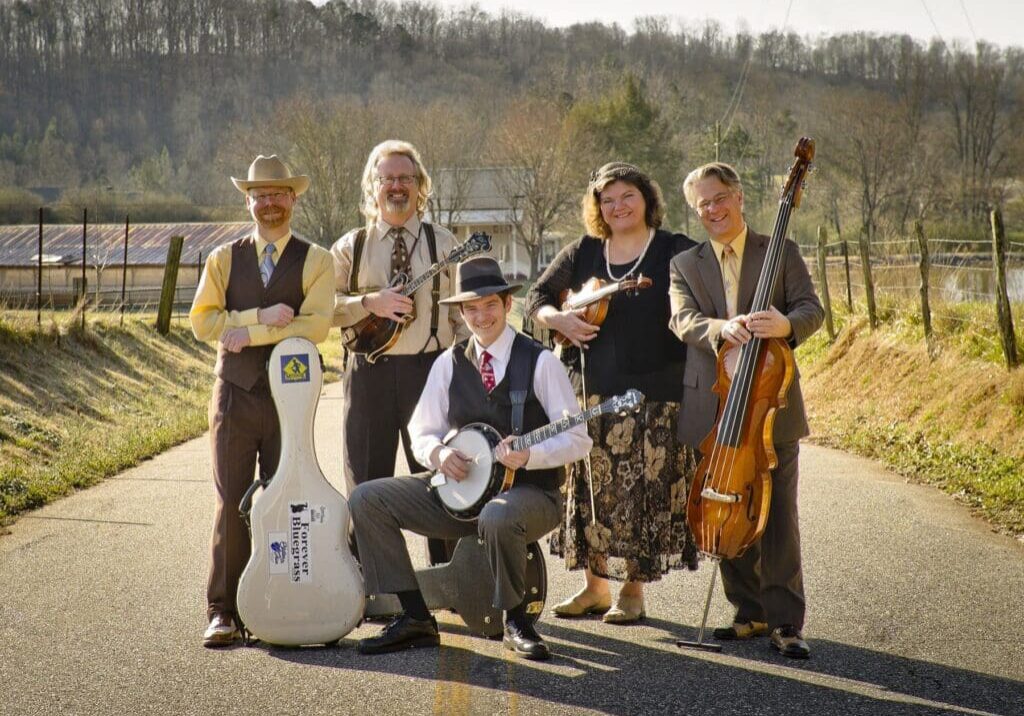 Five musicians pose on a rural road with their instruments and vintage-style outfits, smiling amid trees and a barn—capturing the charm of early spring events in Alexandria, MN.