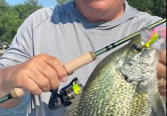 A smiling man in sunglasses and a bright cap holds a fishing rod and proudly displays a large crappie he caught while fishing, standing by the water with trees in the background.