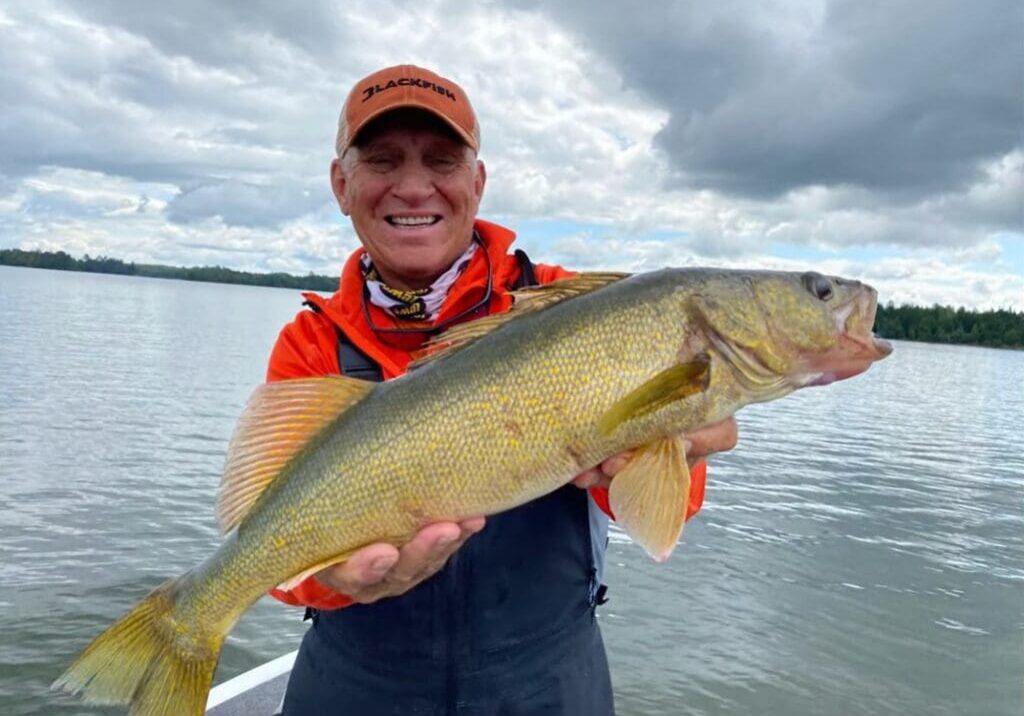 A smiling person in an orange hat and jacket holds a large fish by a lake, proudly showing off their fishing rig, with water and cloudy sky in the background.