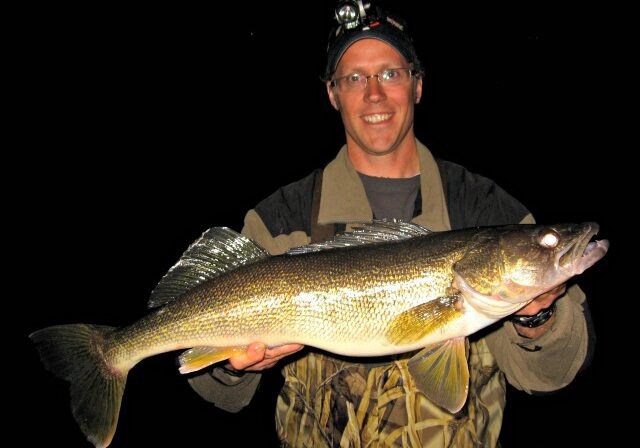 A person wearing glasses and a headlamp smiles while holding a large, shiny-scaled fish, posing against a dark background—just the kind of catch you’d expect to see in an Alexandria May Fishing Report.