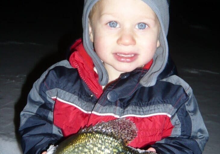 A young child in a red and gray winter jacket and hat proudly holds a large fish with both hands while standing outside in the snow at night, capturing the excitement of a Christmas Ice Fishing Report.