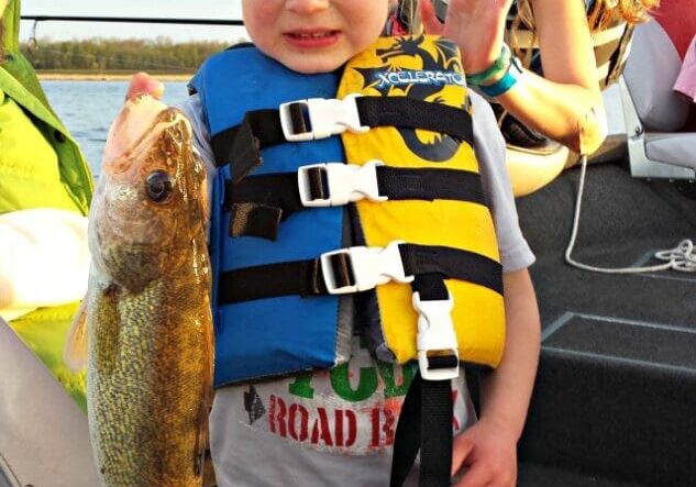 A young boy wearing a blue and yellow life jacket proudly holds a large fish on a boat, while a smiling girl in the background flashes a "four" hand sign and poses cheerfully—capturing the spirit of an early summer fishing report for Alexandria.
