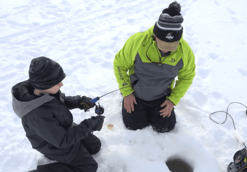Two people dressed in winter clothing kneel on snow next to an ice-fishing hole. One person holds a small fishing rod while the other watches, enjoying a peaceful fishing moment. Ice fishing equipment is nearby.
