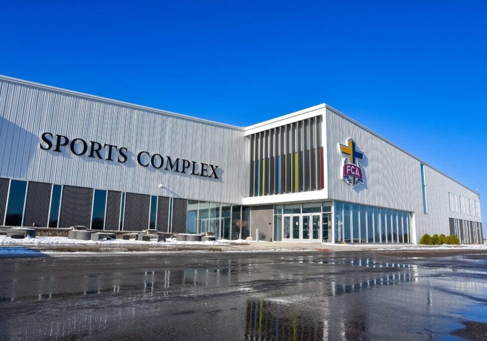 A modern NorthStar Sports Complex building with large vertical windows and "SPORTS COMPLEX" and the "FCA" logo on the exterior. Snow and wet pavement are visible in front under a clear blue sky.