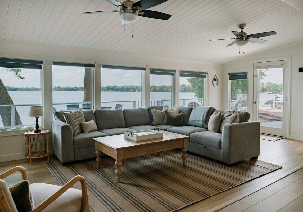 Bright living room at Alex Landing with large windows overlooking a lake, featuring a gray sectional sofa with cushions, a wooden coffee table on a striped rug, ceiling fans, a chair, and a door leading to the deck outside.